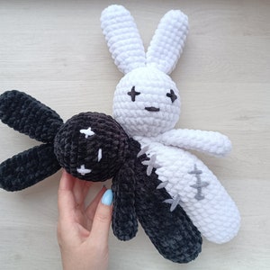 May include: Two crocheted bunny plushies, one black and one white, with stitched eyes and mouths. The black bunny has a white cross on its forehead, and the white bunny has a black cross on its forehead.