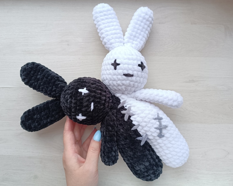 May include: Two crocheted bunny plushies, one black and one white, with stitched eyes and mouths. The black bunny has a white cross on its forehead, and the white bunny has a black cross on its forehead.