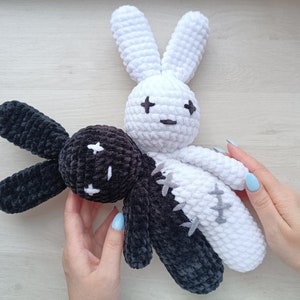 May include: Two crocheted bunny plushies, one black and one white, with stitched eyes and a cross pattern on their bodies. The plushies are joined together, with the black bunny's head resting on the white bunny's shoulder.