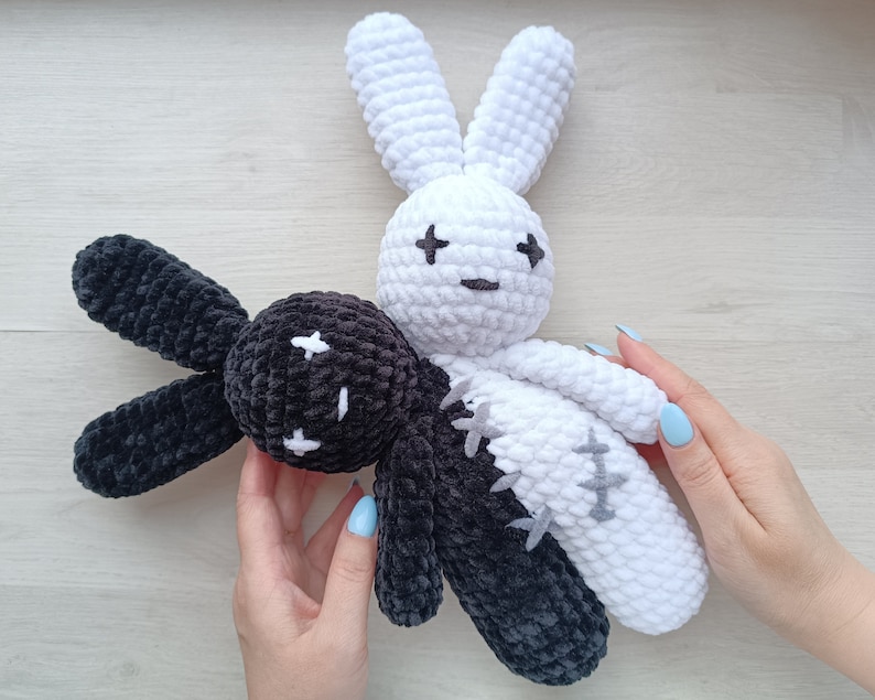 May include: Two crocheted bunny plushies, one black and one white, with stitched eyes and a cross pattern on their bodies. The plushies are joined together, with the black bunny's head resting on the white bunny's shoulder.