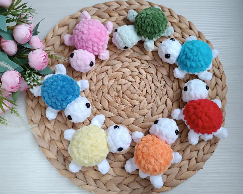 Pu&ograve; includere: Una collezione di otto giocattoli di tartaruga all'uncinetto in vari colori, tra cui rosa, blu, verde, rosso, giallo e arancione. Le tartarughe sono disposte in cerchio su un tappetino di paglia intrecciato.