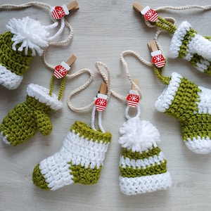 May include: Six crocheted ornaments in the shape of mittens and hats, with white and green stripes. Each ornament is attached to a white string with a small red and white Christmas-themed clip.