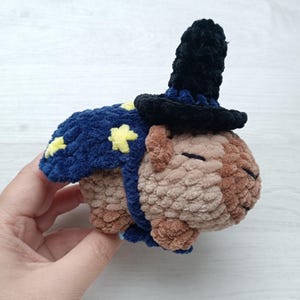 Puede incluir: Un juguete de peluche de capibara hecho de hilo marrón suave, con una capa azul oscuro decorada con estrellas amarillas y un sombrero puntiagudo negro. El capibara tiene los ojos cerrados y una apariencia redondeada y texturizada.