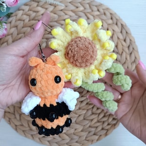 May include: A handmade crochet bee and sunflower. The bee is orange, black, and white with black eyes. The sunflower is yellow, brown, and green. The bee has a black string attached to it.