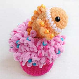 May include: A crocheted orange creature with a white stripe and black eye, adorned with yellow beads, nestled inside a textured pink base featuring loops of yarn and scattered blue and pink beads. The plush toy has a soft, fuzzy, yarn-based construction.