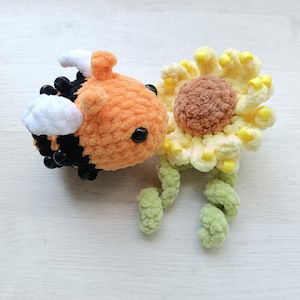 May include: A crocheted plush bee with an orange head, black and orange striped body, and white wings, positioned next to a crocheted sunflower with a brown center, pale yellow petals accented with yellow beads, and two green stems.