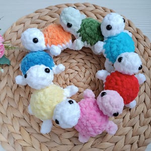 Pu&ograve; includere: Una collezione di giocattoli di tartaruga all'uncinetto in vari colori, tra cui blu, rosso, giallo, rosa, verde e arancione. Le tartarughe sono disposte in cerchio su un cesto intrecciato.