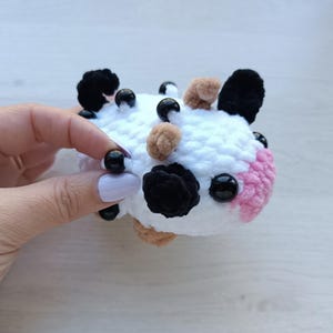 Pode incluir: Um pequeno brinquedo de vaca em crochê branco com manchas pretas e castanhas, olhos de contas pretas e um focinho rosa. A construção em fio macio e texturizado inclui pequenas contas pretas espalhadas pela superfície do objeto.