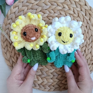 Könnte beinhalten: Zwei handgefertigte gehäkelte Blumenplüschtiere. Eines ist gelb und braun, ähnelt einer Sonnenblume, und das andere ist weiß und gelb, ähnelt einer Gänseblümchen. Beide haben lächelnde Gesichter und grüne Stiele. Die Plüschtiere liegen auf einer gewebten Matte.