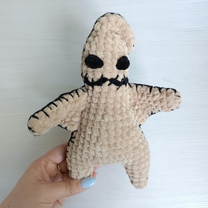 May include: A beige plush toy resembling Oogie Boogie, with black stitched features. The doll has large, black oval eyes and a stitched mouth. The arms and legs are spread out. The doll is held by a hand.