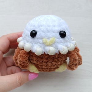 May include: A handmade crochet octopus plushie with a white head, brown body, and black eyes. The plushie has a yellow beak and white bead-like accents around the head. This soft toy is a cute and cuddly item.