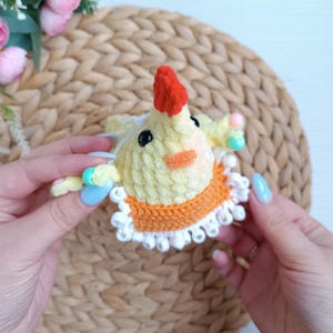 May include: A handmade, yellow crochet chick with a red comb, black eyes, and an orange beak. The chick has a white and orange bib and small wings with blue and pink accents. The chick is a soft, plush toy.