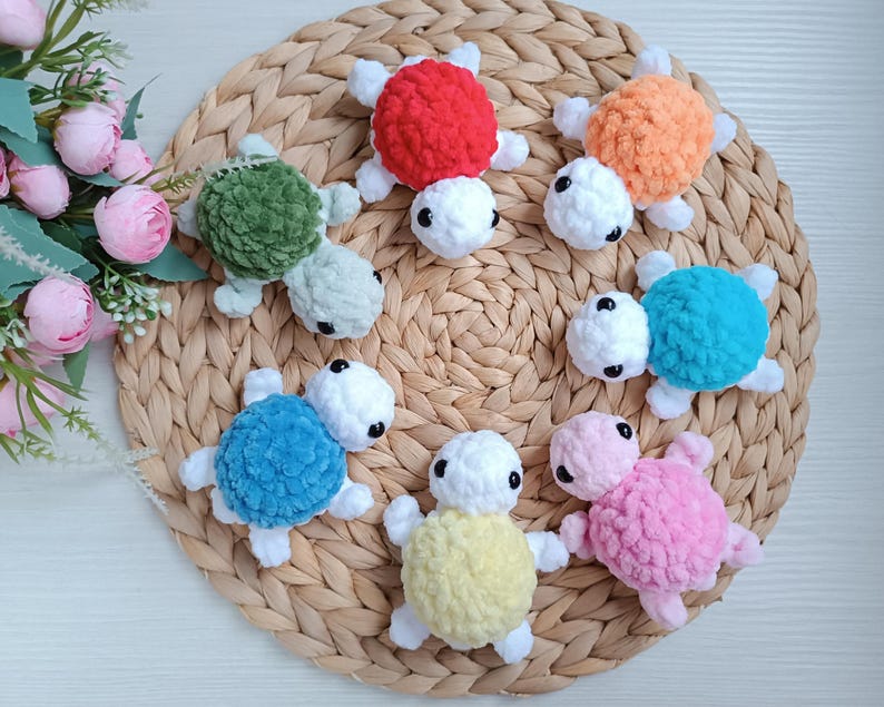 Pu&ograve; includere: Sette giocattoli di tartaruga all'uncinetto in vari colori, tra cui verde, rosso, arancione, blu, rosa e giallo. Le tartarughe sono disposte su un tappetino di paglia intrecciata.
