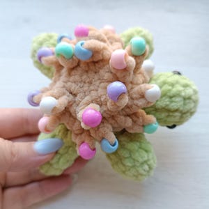 May include: A handmade, plush turtle toy in shades of green and tan. The turtle's body is tan with colourful beads, and the head and flippers are green. The toy is soft and textured, with a whimsical design.