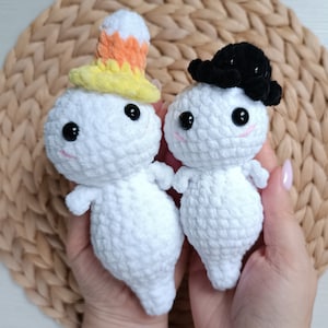 May include: Two white crocheted ghost figures with plush texture. One ghost wears a yellow and orange candy corn hat, while the other wears a black hat. Both figures feature round black eyes and small pink stitched mouths, crafted from soft yarn.