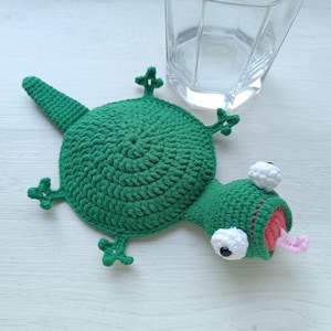 May include: A green crocheted lizard-shaped coaster featuring a round body, four small feet, a long tail, and a head with two white protruding eyes, a red interior mouth, and a light pink tongue sticking out, positioned near a clear glass.