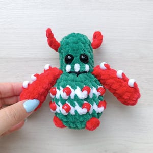May include: A plush monster toy crafted from green and red yarn. The creature features two red horns, large black plastic eyes, and a wide mouth with white teeth. Its body and arms are decorated with small white and red plastic beads throughout.