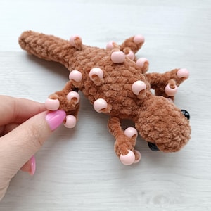 Puede incluir: Un juguete de lagarto de ganchillo marrón hecho a mano con detalles de cuentas rosas. El reptil de peluche tiene ojos negros y está sobre una superficie de color claro. El cuerpo del lagarto está cubierto de pequeñas cuentas rosas redondas.