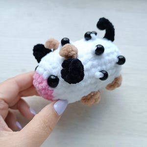 Peut inclure: Un petit jouet en peluche crocheté blanc en forme de vache avec des taches noires et marron clair, un museau rose, des yeux en perles noires et plusieurs perles noires fixées sur le dos. La peluche a une petite queue noire et des pieds crochetés marron clair.
