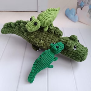 Pode incluir: Três brinquedos de crocodilo verde de crochê. O crocodilo maior está deitado com a cabeça virada para o lado. Um crocodilo menor está sentado em cima do crocodilo maior. O crocodilo menor está em pé na superfície branca.