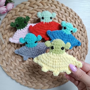 Può includere: Una collezione di peluche alieni fatti a mano all'uncinetto in vari colori, tra cui verde, rosso, blu e giallo. Ogni peluche ha una testa rotonda con piccole orecchie e occhi neri. I giocattoli sono disposti su un tappetino intrecciato.