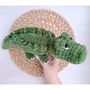 May include: A handmade green plush crocodile toy with white claws and black eyes. The crocodile is made of a soft, textured material and is lying on a woven, circular mat. The toy is approximately 30 cm long.
