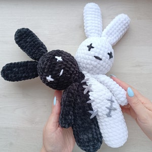 May include: Two crocheted bunny plushies, one black and one white, are joined together at the torso. The black bunny has a stitched-on face with black eyes and a stitched-on mouth. The white bunny has a stitched-on face with black eyes and a stitched-on mouth. The bunnies are held by a hand.