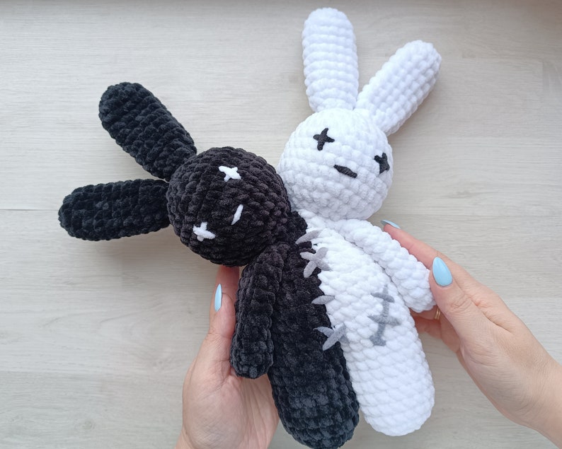May include: Two crocheted bunny plushies, one black and one white, are joined together at the torso. The black bunny has a stitched-on face with black eyes and a stitched-on mouth. The white bunny has a stitched-on face with black eyes and a stitched-on mouth. The bunnies are held by a hand.