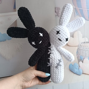 May include: Two crocheted bunny plushies, one black and one white, are sewn together to create a yin yang design. The plushies have embroidered eyes and stitched mouths.