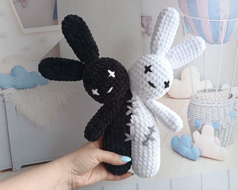 May include: Two crocheted bunny plushies, one black and one white, are sewn together to create a yin yang design. The plushies have embroidered eyes and stitched mouths.