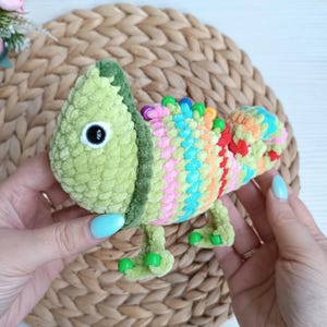 Puede incluir: Un juguete de camaleón de ganchillo hecho a mano en tonos verdes, rosas, naranjas y azules. El camaleón tiene un cuerpo texturizado y difuso, ojos negros grandes y pequeños pies verdes. La espalda del juguete presenta un patrón de rayas en colores arcoíris.