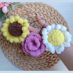 May include: Three handmade flower hair accessories. A yellow sunflower with a brown center, a pink rose, and a white daisy with a yellow center. The flowers are made of a soft, textured yarn and are attached to black elastic hair ties.