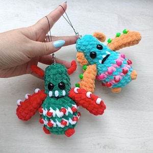 May include: Two plush monster charms made of soft yarn. One is dark green with red arms, white teeth, and red beads. The other is turquoise with orange arms, black eyes, and multi-colored beads. Both feature black string loops for hanging as accessories.