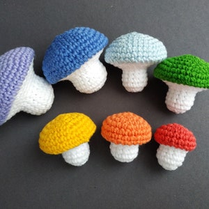 K&ouml;nnte beinhalten: Ein Set aus acht geh&auml;kelten Pilzen in verschiedenen Farben, darunter Lila, Blau, Hellblau, Gr&uuml;n, Gelb, Orange und Rot. Die Pilze sind alle mit einer wei&szlig;en Basis gefertigt und haben eine geh&auml;kelte Kappe in verschiedenen Farben.
