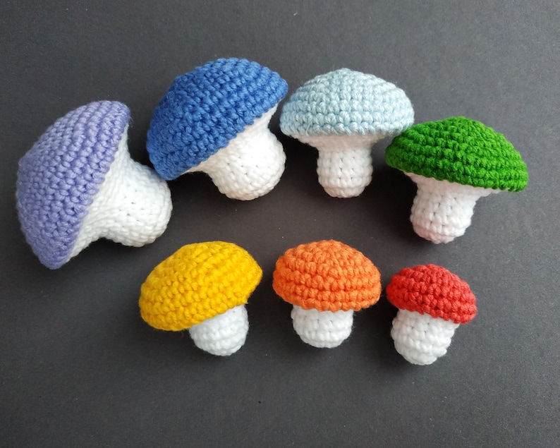 K&ouml;nnte beinhalten: Ein Set aus acht geh&auml;kelten Pilzen in verschiedenen Farben, darunter Lila, Blau, Hellblau, Gr&uuml;n, Gelb, Orange und Rot. Die Pilze sind alle mit einer wei&szlig;en Basis gefertigt und haben eine geh&auml;kelte Kappe in verschiedenen Farben.