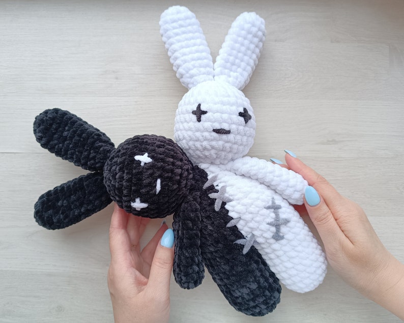 May include: Two crocheted bunny plushies, one black and one white, with stitched eyes and a cross pattern on their bodies. The plushies are joined together, with the black bunny's head resting on the white bunny's shoulder.