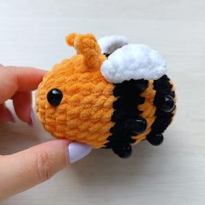 May include: A small, plush, crocheted bee toy featuring a rounded body with alternating horizontal stripes of bright orange and black yarn. The bee has two soft white wings on its back, two small antennae on top, and two shiny black plastic bead eyes.