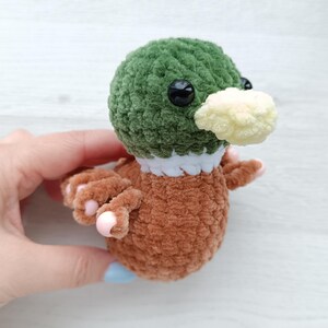 May include: A handmade crochet duck toy. The duck has a green head, brown body, and a cream-colored beak. It has black safety eyes and small pink claws. The duck is made of soft, plush yarn.