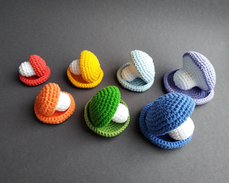 K&ouml;nnte beinhalten: Ein Set aus sieben geh&auml;kelten Muscheln in verschiedenen Farben, darunter Rot, Orange, Gelb, Gr&uuml;n, Blau und Lila. Jede Muschel enth&auml;lt eine wei&szlig;e Perle.