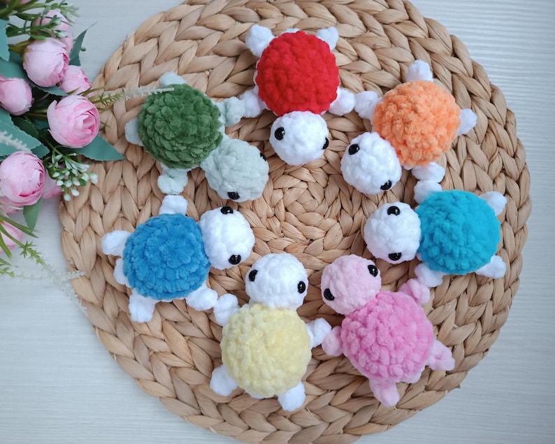 Pu&ograve; includere: Sette giocattoli di tartaruga all'uncinetto in diversi colori, tra cui rosso, arancione, verde, blu, giallo, rosa e bianco. Le tartarughe sono disposte in cerchio su una superficie di vimini intrecciato.
