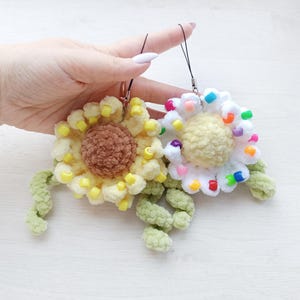 Könnte beinhalten: Zwei handgefertigte gehäkelte Sonnenblumen-Anhänger. Einer ist gelb mit brauner Mitte und gelben Perlen, der andere ist weiß mit gelber Mitte und bunten Perlen. Beide haben grüne Stiele und schwarze Lanyards.