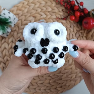 May include: A handmade white crochet owl with large black eyes and black bead accents. The owl has a fluffy texture and is held in a person's hands. The owl's beak is black, and the owl is a decorative item.
