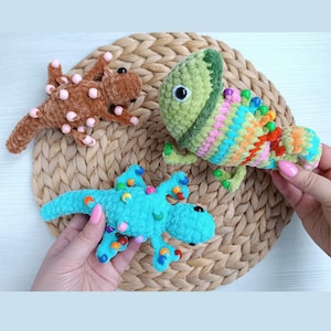 May include: Three crocheted lizard toys in brown, green, and blue, each with colorful bead accents. The brown lizard has pink beads, the green one has a rainbow of colors, and the blue one has orange, blue, and green beads. The toys are on a woven mat.