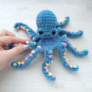 Puede incluir: Un pulpo azul tejido a ganchillo con ojos negros y cuentas de colores en sus tentáculos. El peluche está hecho de hilo suave y texturizado. El pulpo es un artículo hecho a mano.