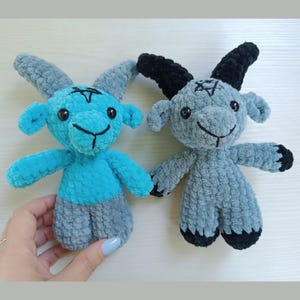 May include: Two crocheted goat plushies. One is blue and gray, the other is gray and black. Both have horns, a pentagram on their foreheads, and smiling faces. The plushies are handmade and have a soft, textured appearance.