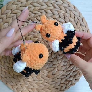 May include: Two handmade crochet bee plushies featuring soft orange yarn bodies with black stripes and white wings. Each bee has small black plastic eyes and is attached to a thin black cord loop for hanging as a decorative charm or keychain accessory.