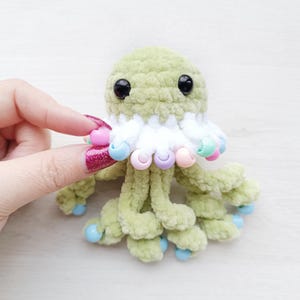 May include: A handmade, light green crochet octopus with black eyes and a white frill. The octopus has multiple tentacles with light blue tips and is adorned with colourful beads. A whimsical, soft toy.