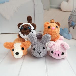 May include: Five crocheted stuffed toys: a brown and white cat, an orange tabby cat, a grey bunny, an orange and white corgi, and a pink pig. The toys are all sitting on a white wooden surface.