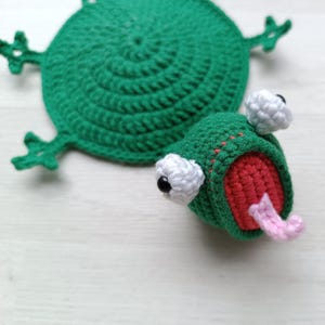 Può includere: Un sottobicchiere all'uncinetto verde a forma di rana con un corpo circolare piatto e quattro piccoli arti. Al corpo è attaccata una testa di rana tridimensionale con occhi bianchi, pupille nere, una bocca rossa aperta e una lingua rosa.