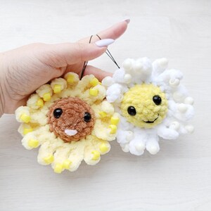 May include: Two handmade crochet flower ornaments. One is yellow with a brown centre and yellow accents, the other is white with a yellow centre and white accents. Both have black eyes and smiling faces. Each has a black string for hanging.